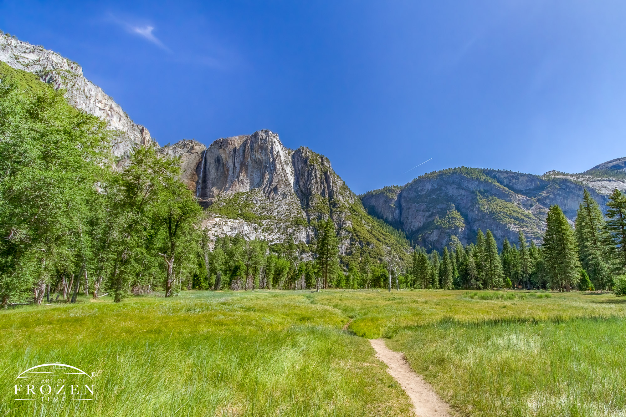 A footpath leads the eye through a grassy meadow with Yosemite Point and Falls on the distant horizon.