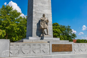 Stone frontiersman sculpture at the base of the Fort Recovery State Memorial in Ohio, with flintlock musket, memorial plaque, officer names, and blue sky.