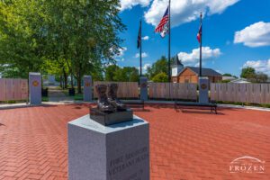 Side view of bronze combat boots at Fort Recovery Veterans Plaza in Ohio, with U.S. flag, Ohio flag, POW-MIA flag, and a sunlit church in the background.