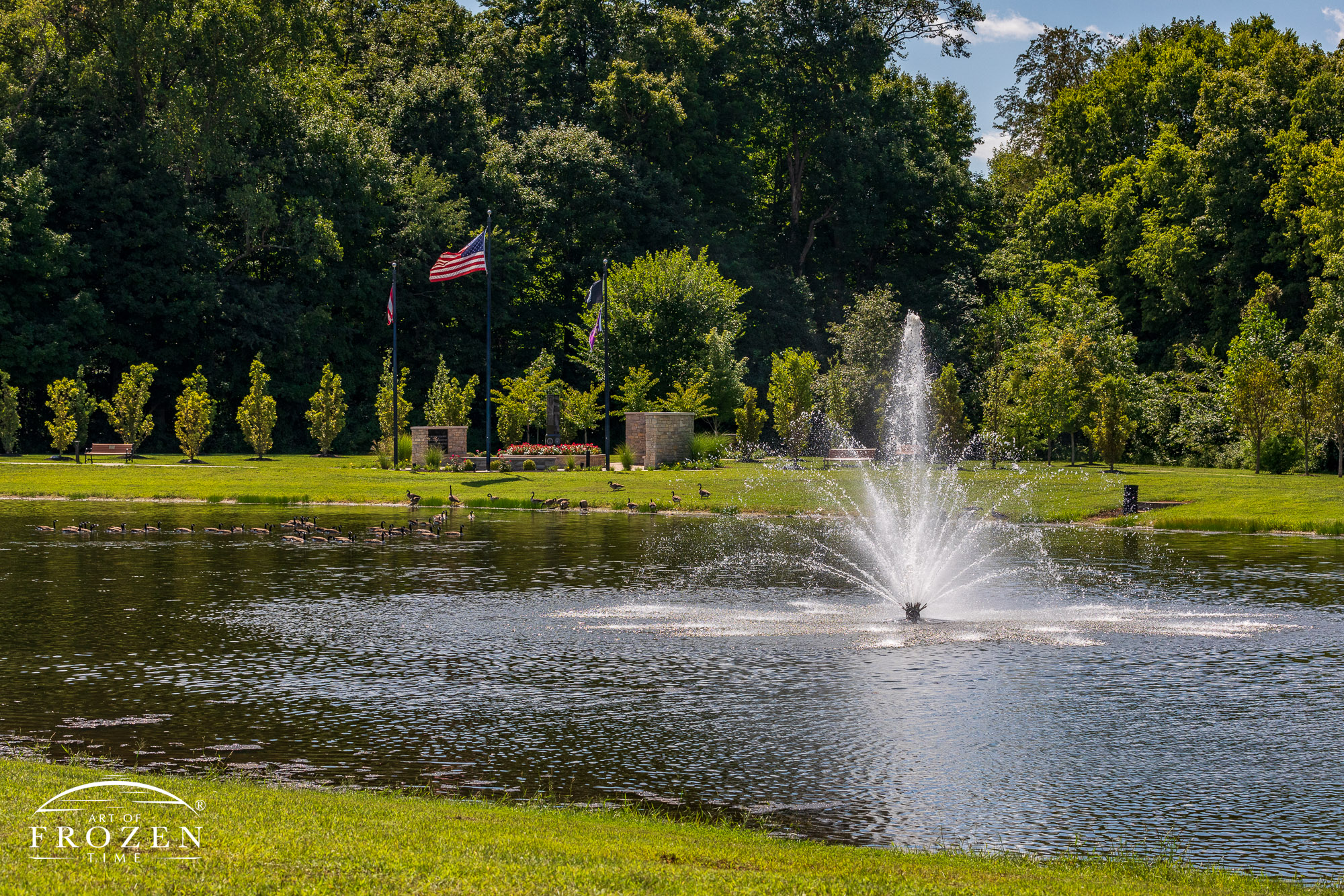 A view of the Gardner Park walking path which traverse the large pond featuring a fountain as the pathway meanders through trees and open space.