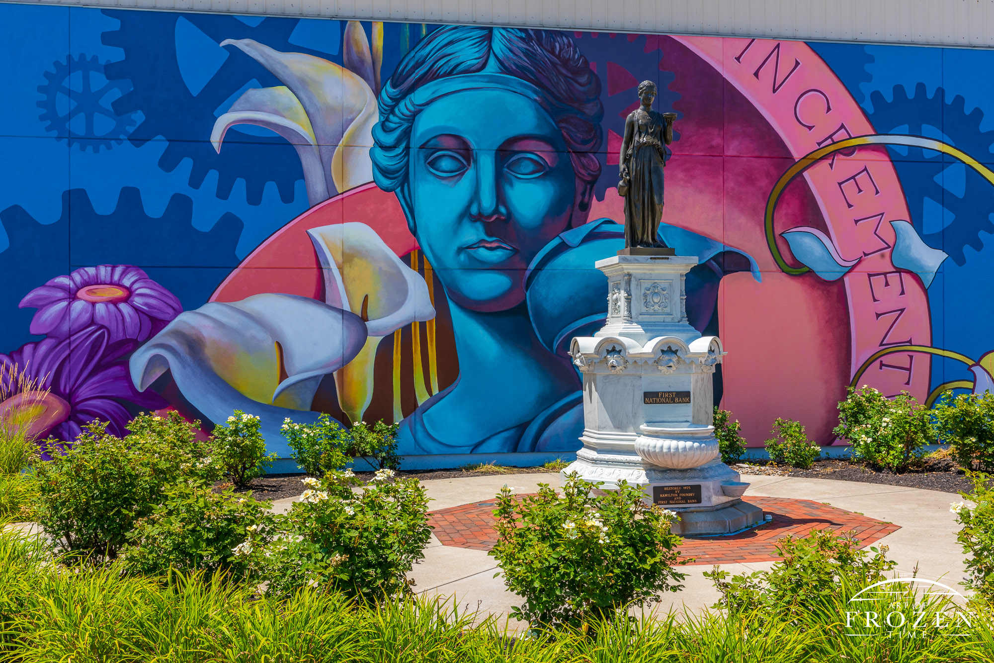 A colorful mural in Hamilton Ohio where the Greek subject of the mural matches the bronze sculpture mounted on a stone plinth.