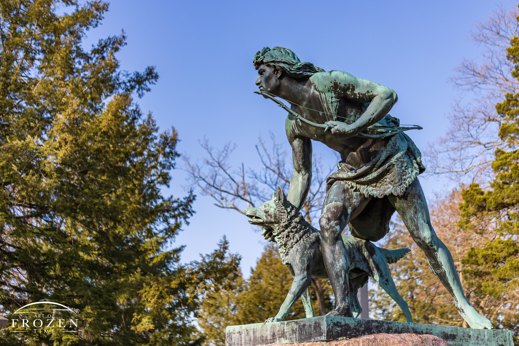 A bronze statue of a native American holding a bow with one hand while steading his snarling dog with the other as they face an unknown encounter