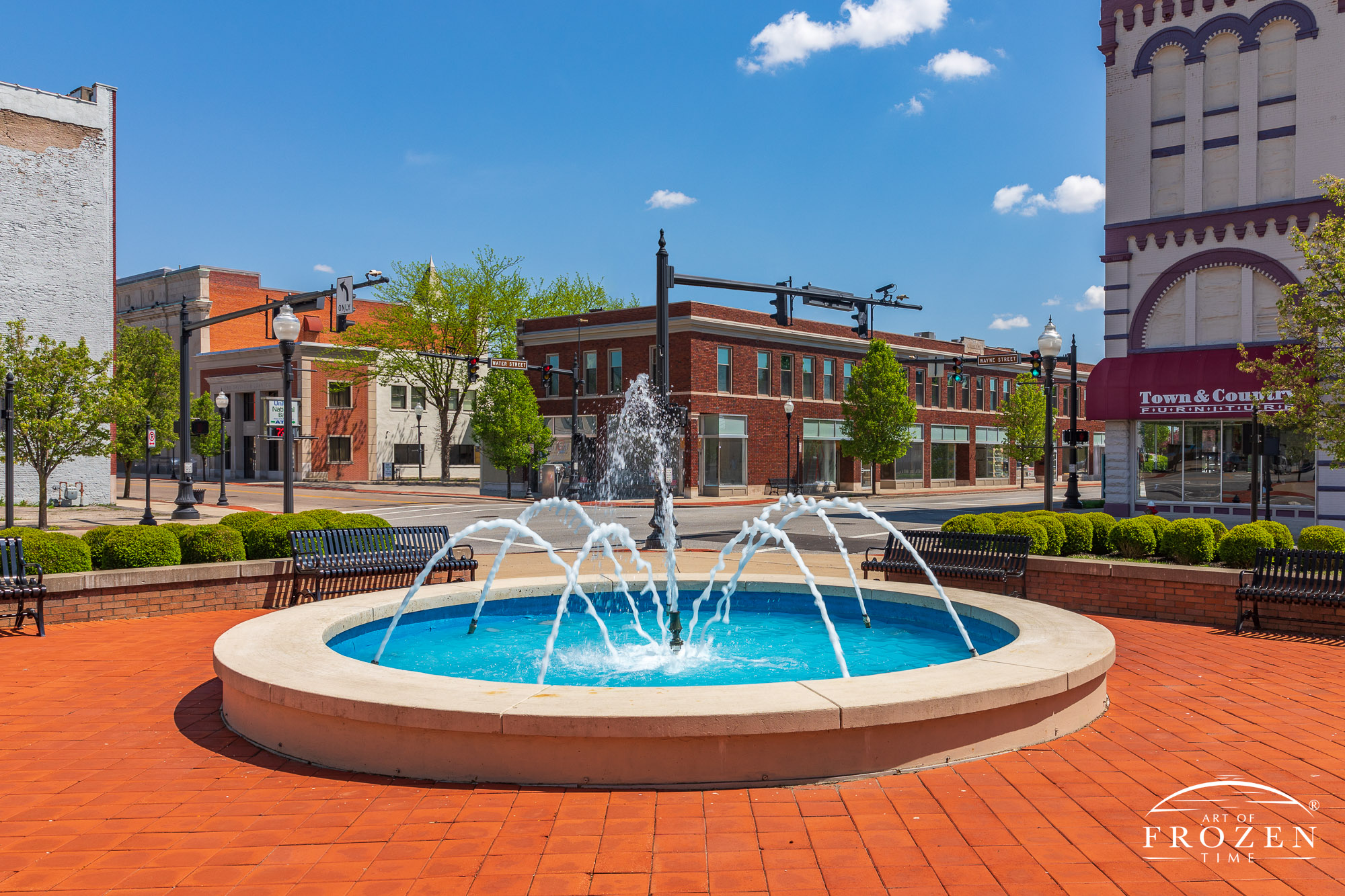 A red paver plaza surrounds a fountain in front of Piqua’s City office building which basks in bright light under a cloudless sky