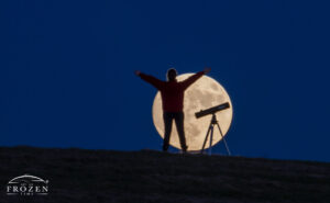 A telephoto view of a girl standing in front of a rising full moon that reveals her silhoutte and telescope