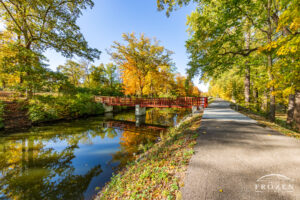A tree-lined paved trail parallels Piqua Ohio’s Hydraulic Canal Run in autumn where the sun illuminated the autumn colored leaves