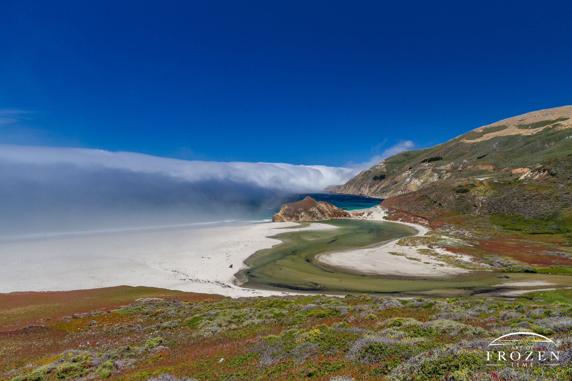 View from California State Route 1 along the Santa Lucia Mountains on a clear blue day with sea fog making its way onshore and the Little Sur River meandering towards the coast