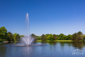 A large fountain in a small tree-lined lake where the vegetation takes on vibrant green colors which complement the impressively blue sky.