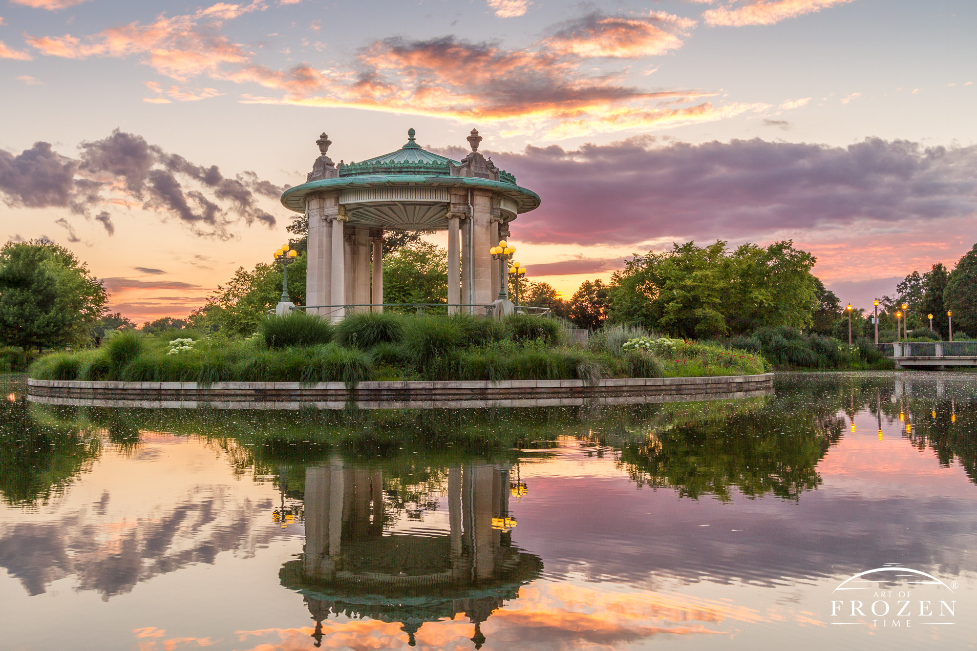A colorful sunset over St Louis Forest Park, where pink and purple clouds hang over the Nathan Frank Bandstand while Pagoda Lake reflects the impressive sky