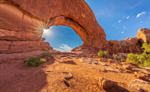 A morning view of the North Windows Arch in Arches National Park where the sun is rendered as a starburst illuminated boulders and brush.