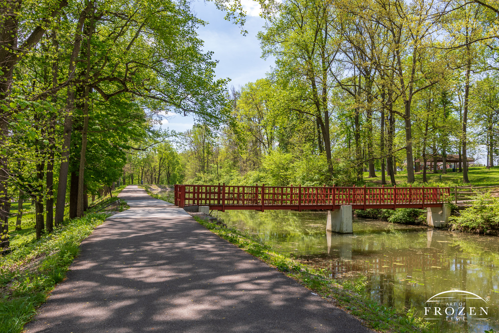 A decorative foot bridge spans Piqua Ohio’s Hydraulic Canal Run as bikers enjoy cool shade from the tree-lined pathway