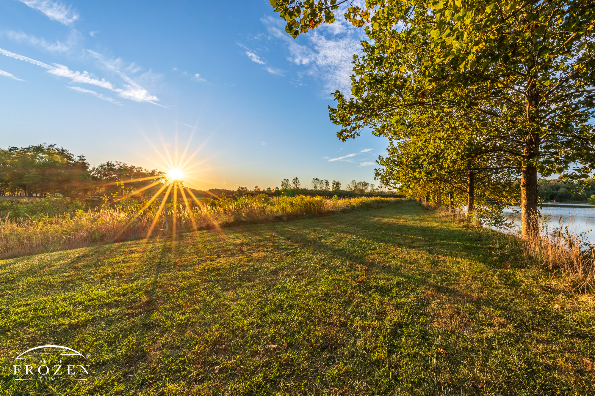 A wide hiking trail through a meadow along the Great Miami River, where the setting sun cast a warm light against the green vegetation.