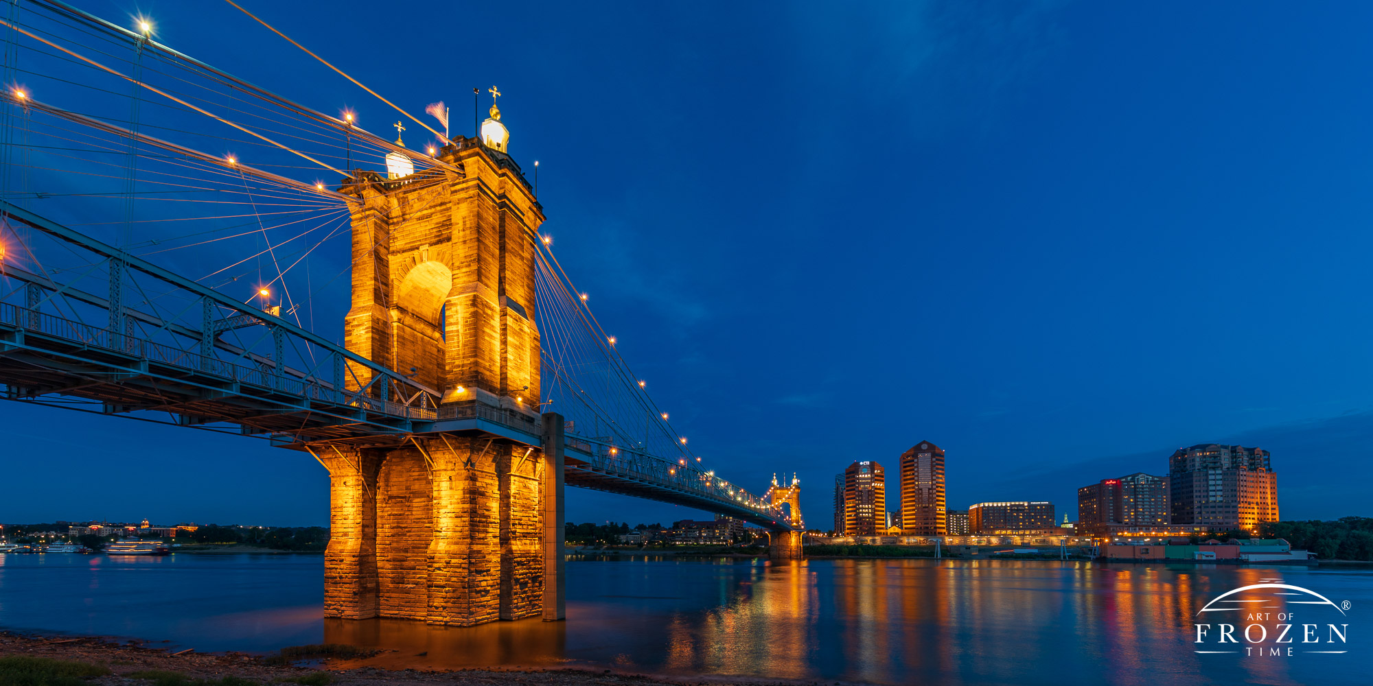 A twilight image of the John A. Roebling Suspension Bridge as its warm yellowish lights illuminates the evening as it spans the Ohio River