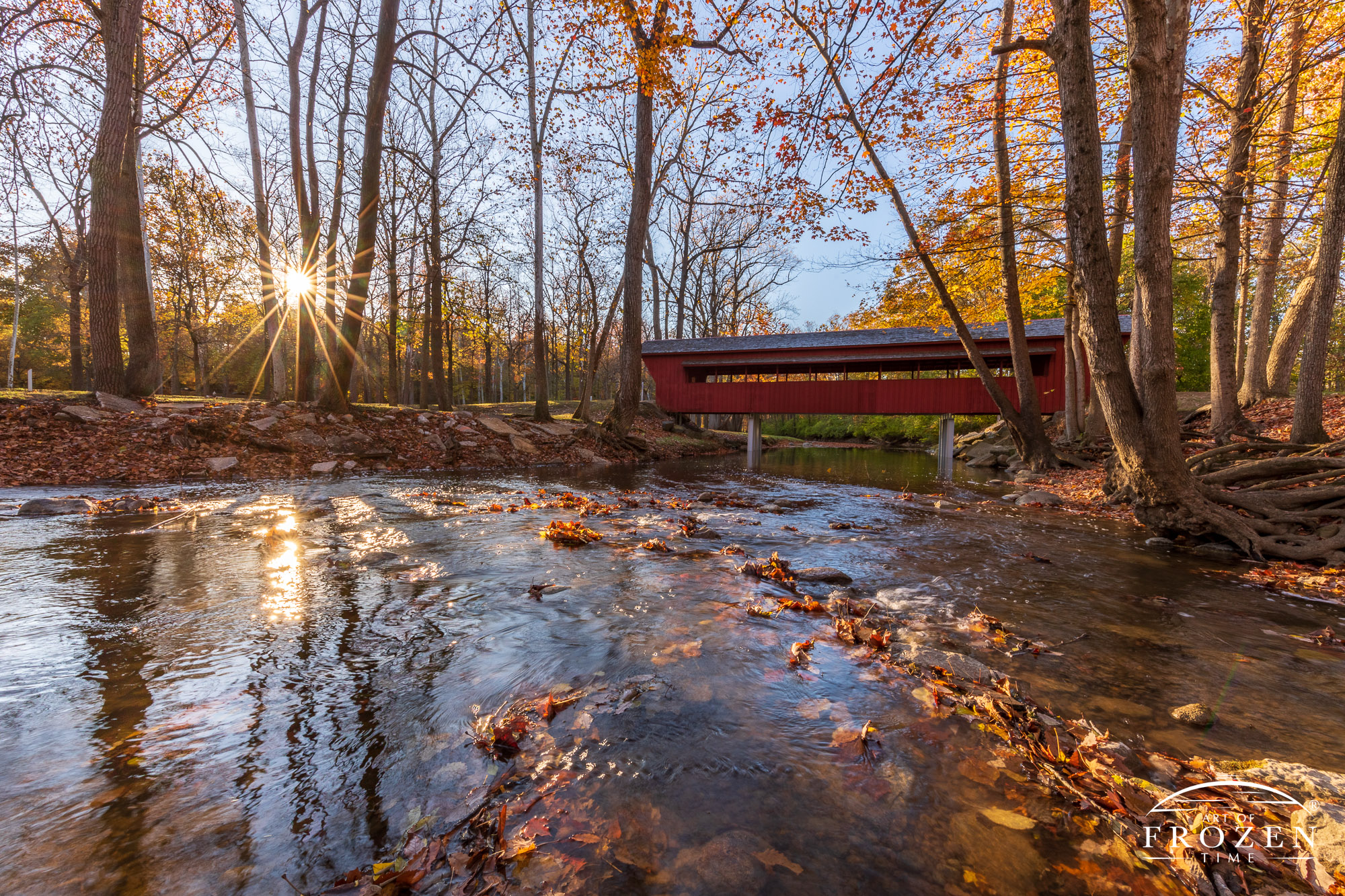 A red wooden bridged spans Tawawa Creek on a colorful autumn evening as the thinning leave barely block the setting sun and its golden light