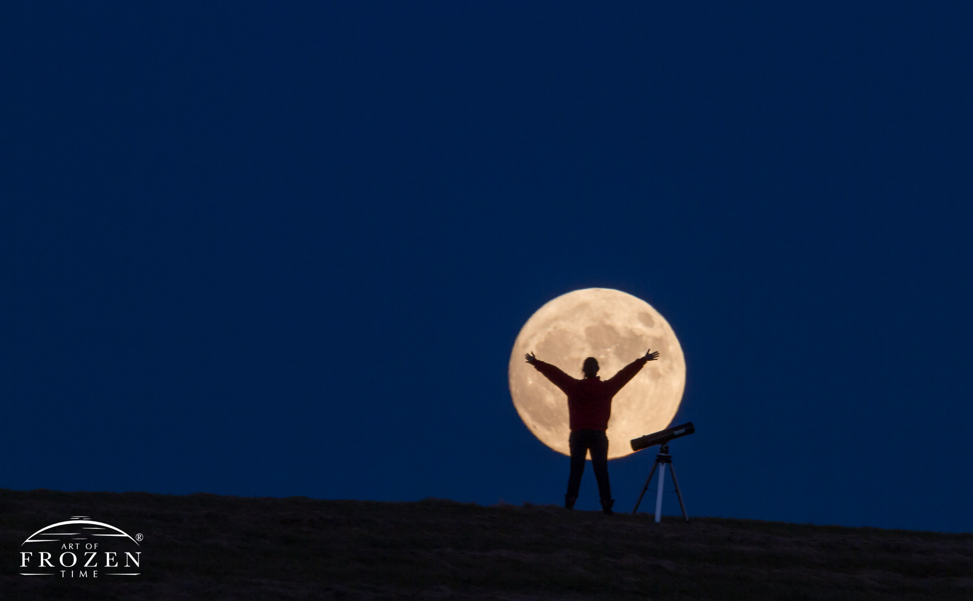 A telephoto view of a girl in front of a giant full moon where the silhoutte reveals her outstretched arms and a nearby telescope