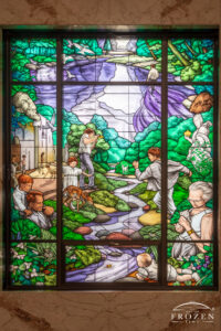 Historic stained-glass windows created by Louis Comfort Tiffany featuring his opalescent glass featuring green and purple colors as a stream runs down the middle as others does things that bring them joy.