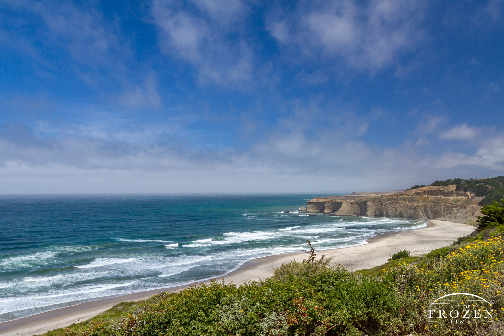 A Highway 1 vista along the California coast revealing a long sandy beach with a fog bank making its way ashore.