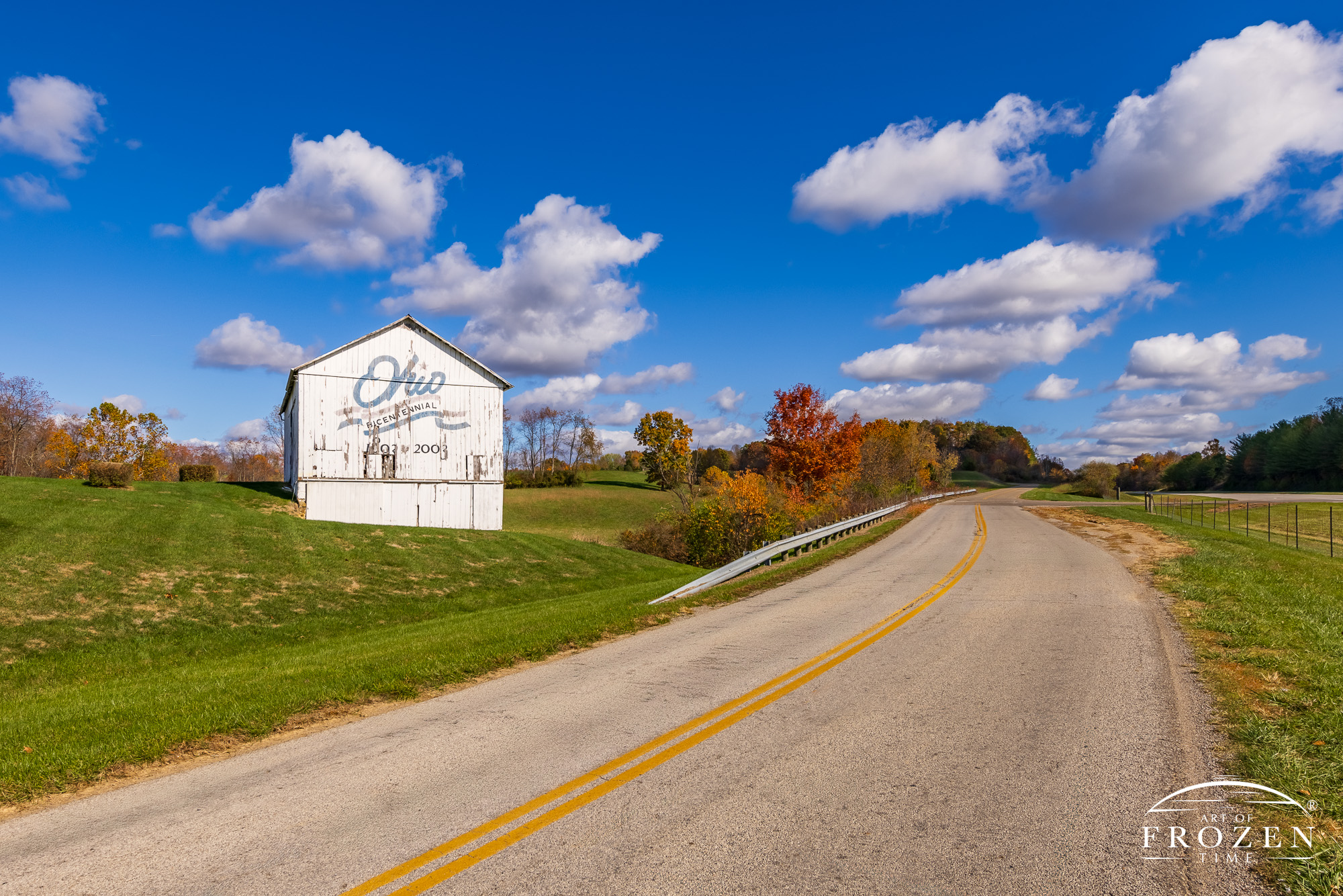An old white barn as its stands among the rolling hills of Athens County displaying the Ohio Bicentennial logo where the paint is fading after 25 years of wear.