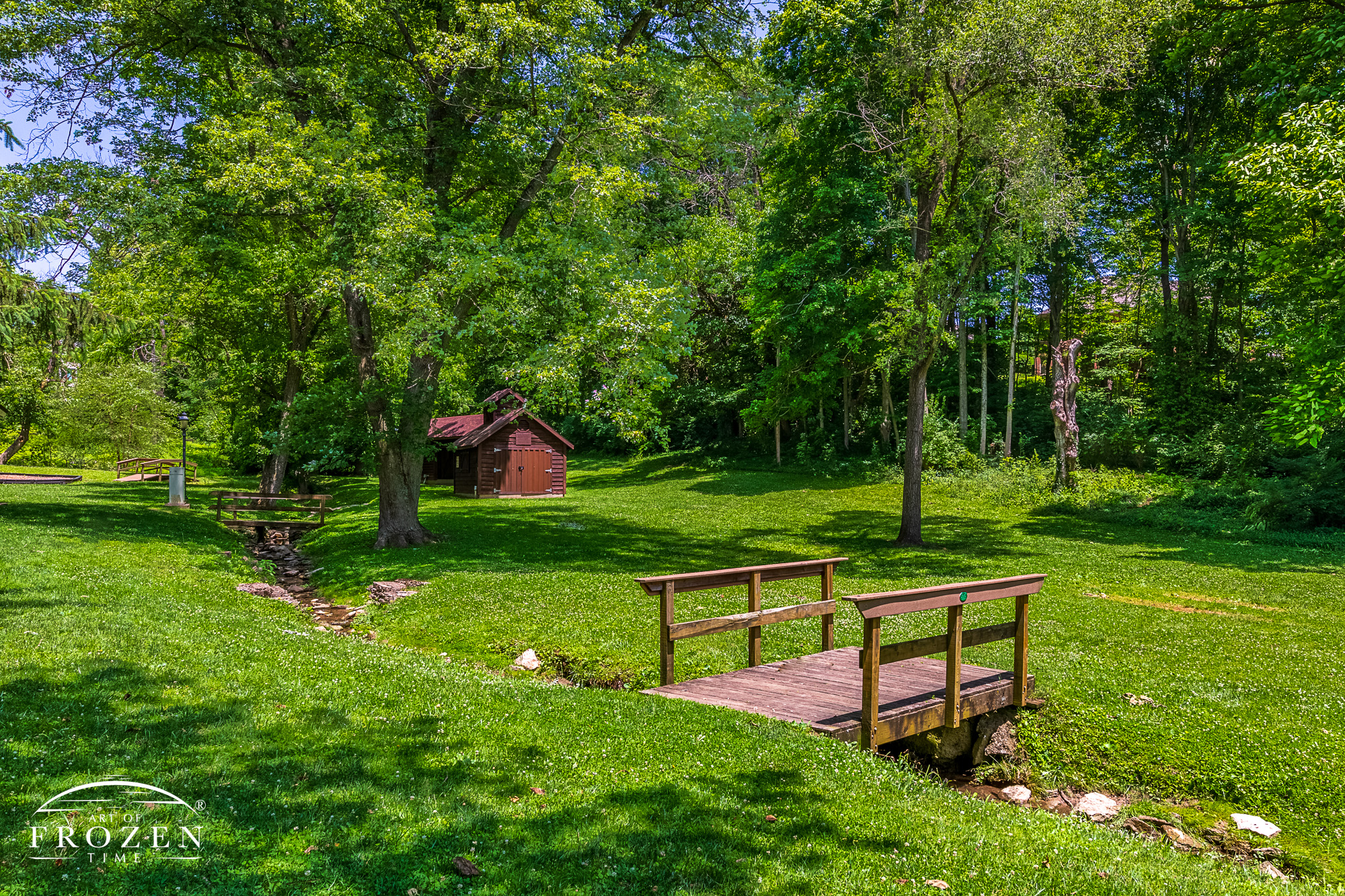 Bellbrook Park in Bellbrook Ohio on a pretty summer day where a foot bridge spans a creek that cuts through the park and leads the eye towards the park's sugar maple house