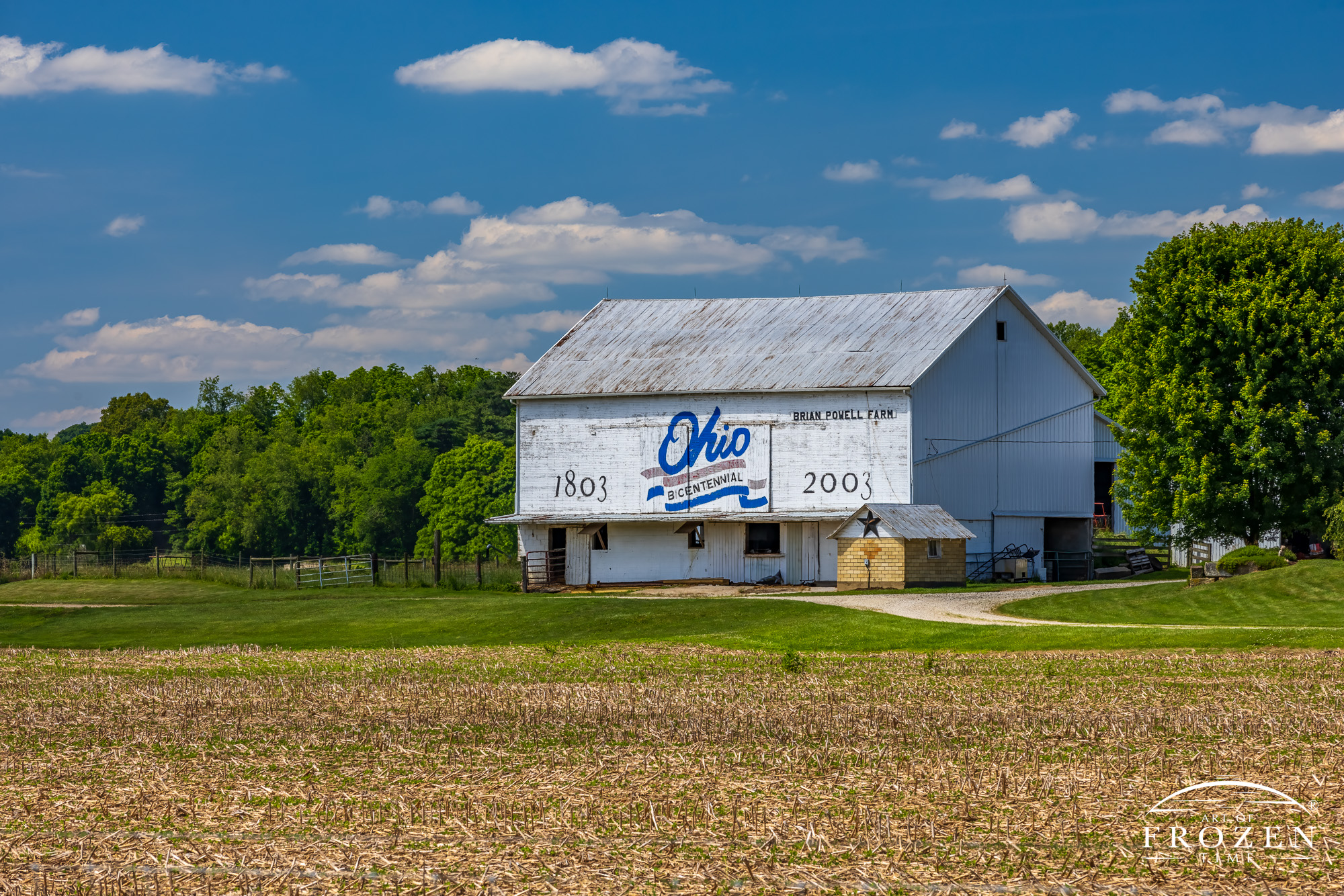 A white barn adorned with the Ohio Bicentennial Logo depicting a scene where the Coshocton barn basks in the summer sun will cumulus clouds floating in the sky
