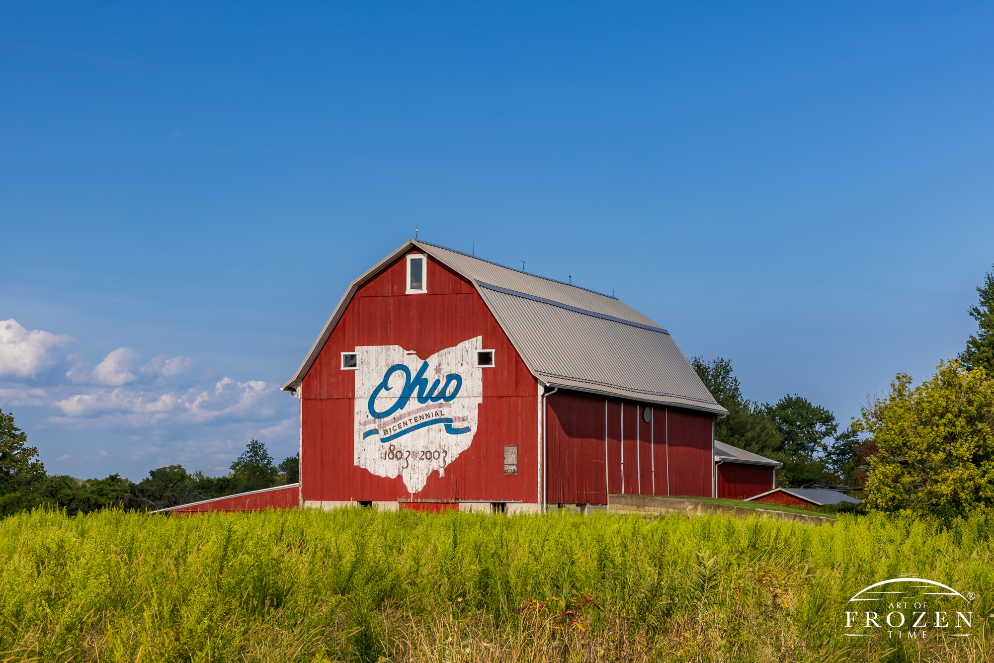 An Ohio landmark art print where the red, Erie County Bicentennial Barn faces the western sky as the evening light paints the red, white and blue logo in warm light. Caption: At the junction of U.S. Route 6 and Perkins Avenue, just four miles east of Sandusky,