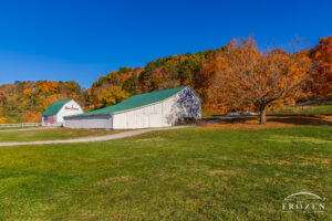 White barn with a green roof and an Ohio sign on the front, set in a park-like field with colorful autumn trees and a clear blue sky.