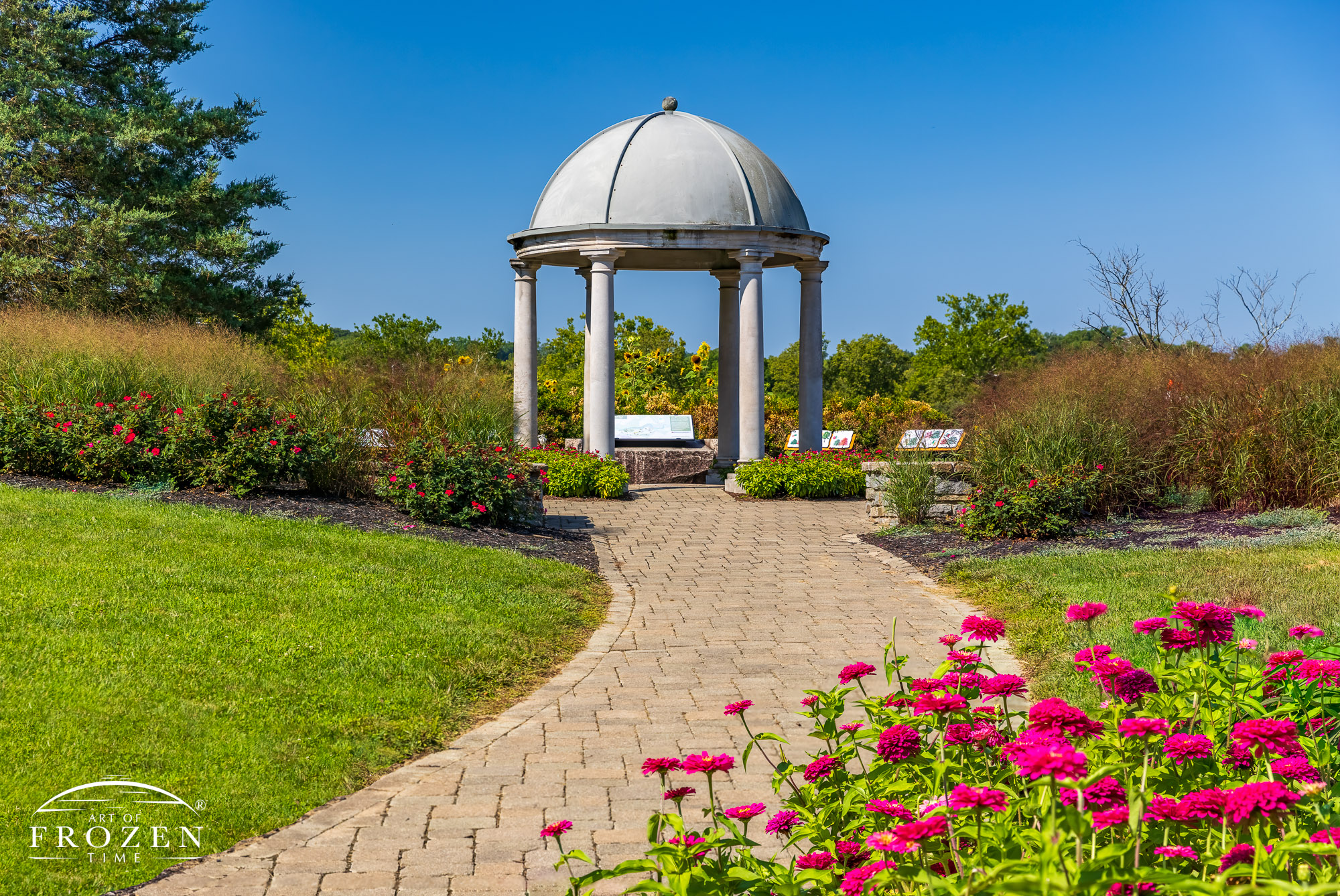 A summer garden view where a paver pathway leads the towards a pretty stone gazebo surrounded by flowers