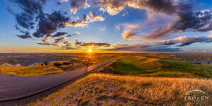 Panoramic sunset over South Dakota Route 240 winding through Badlands National Park with golden light, prairie grass, and dramatic summer clouds