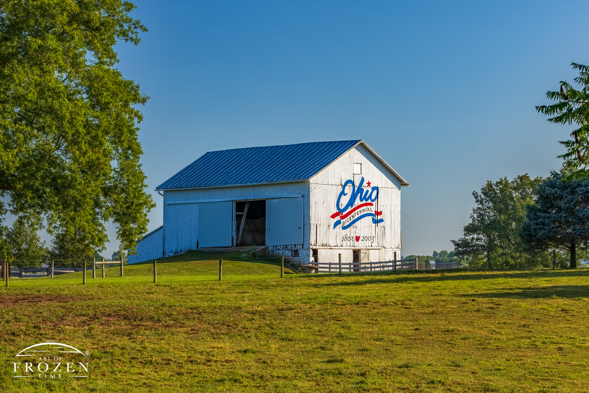 An ancient but sturdy white barn in the morning light as the sun illuminates the patriotic red, white and blue logo painted on its eastern side contrast nicely with the surrounding and lush green pasture
