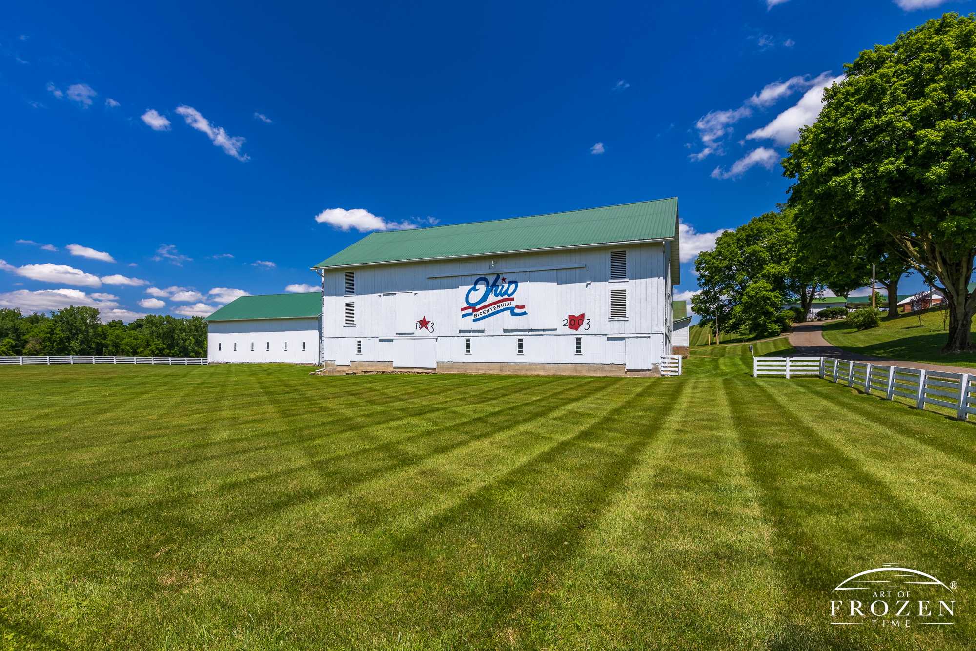 A Holmes County Ohio fine art print of a well maintained white barn featuring the patriotic Ohio Bicentennial Logo where the grounds are perfectly groomed as cumulous clouds float through the blue sky.