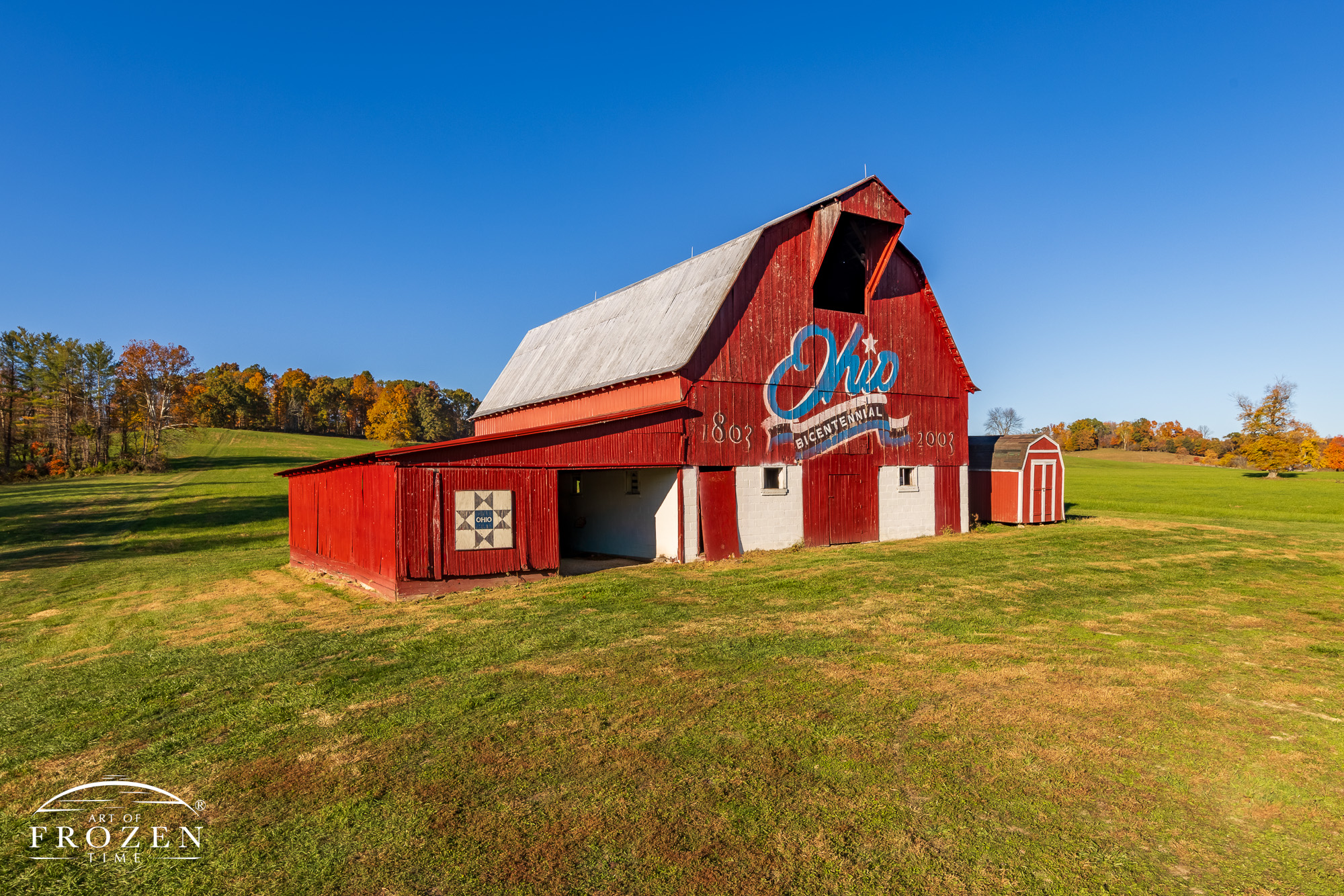 A gorgeous red barn displaying a blue version of the Ohio Bicentennial logo on an autumn evening where the warm light paints the barn in surrounding hillside in perfect light.