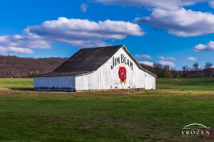 White Jim Beam barn at the James B. Beam Distilling Co. in Clermont, Kentucky, with spring grass, rolling hills, blue sky, and broken sunlight.