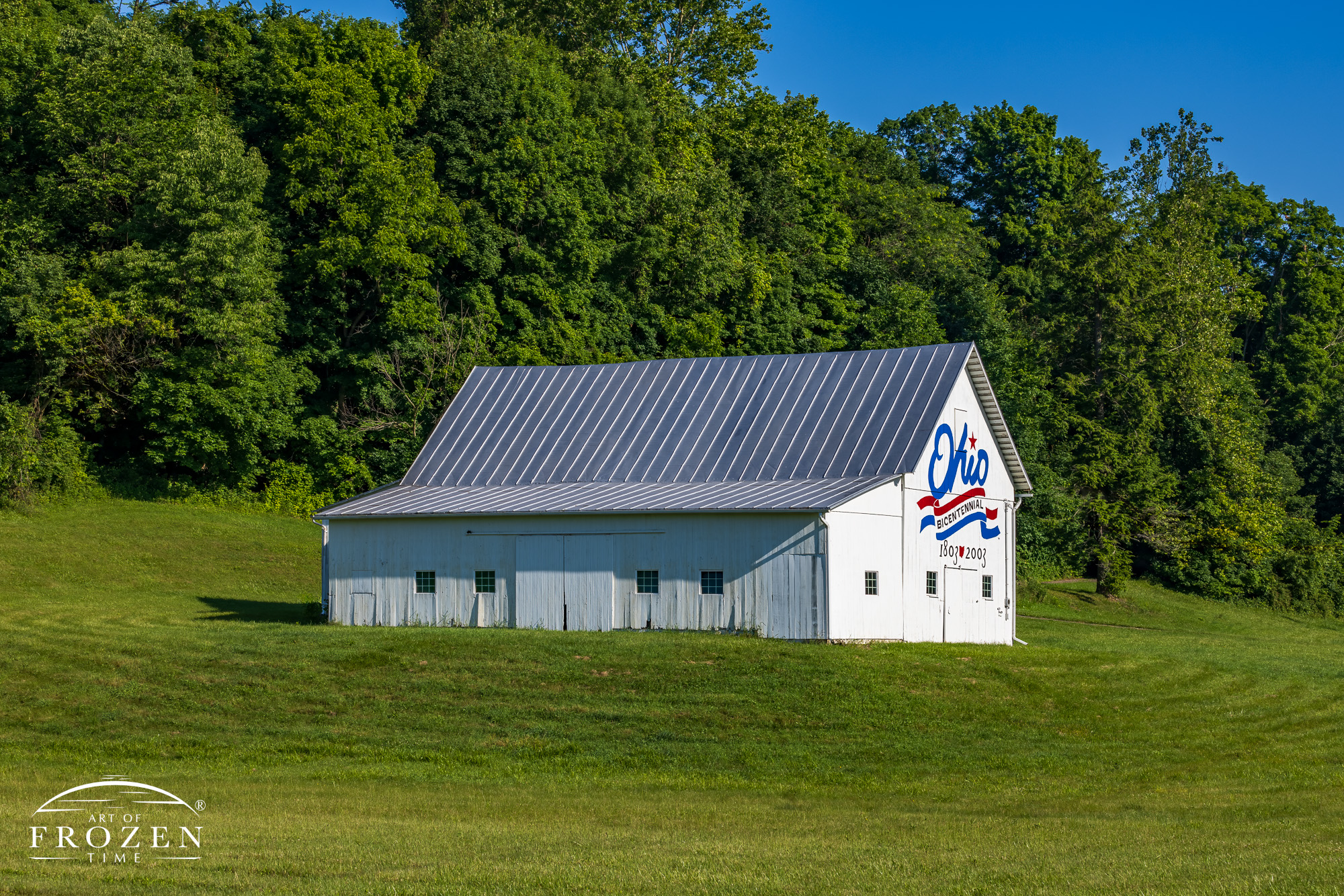 An Ohio favored landmark entails the Licking County Ohio Bicentennial Barn where its bright white paint contrast with the lush green hillside as the logo basks in the summer morning light