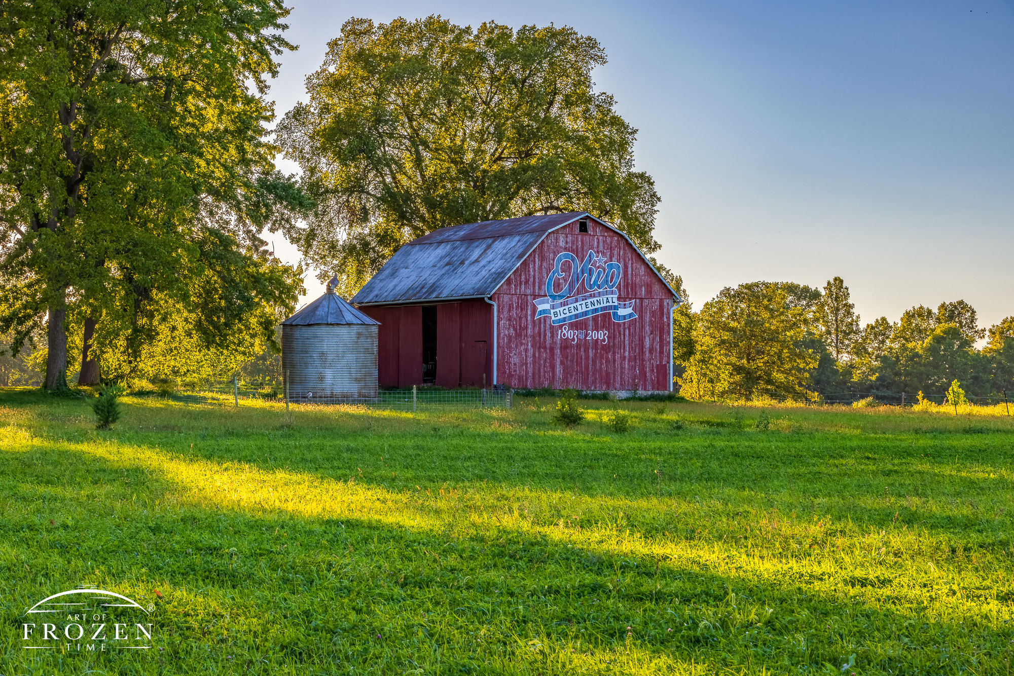 An old Ohio red barn from a distance that serves as the Logan County Ohio Bicentennial Barn. During this evening shot, the surrounding trees gently filter the warm light as it rakes across the adjacent field.