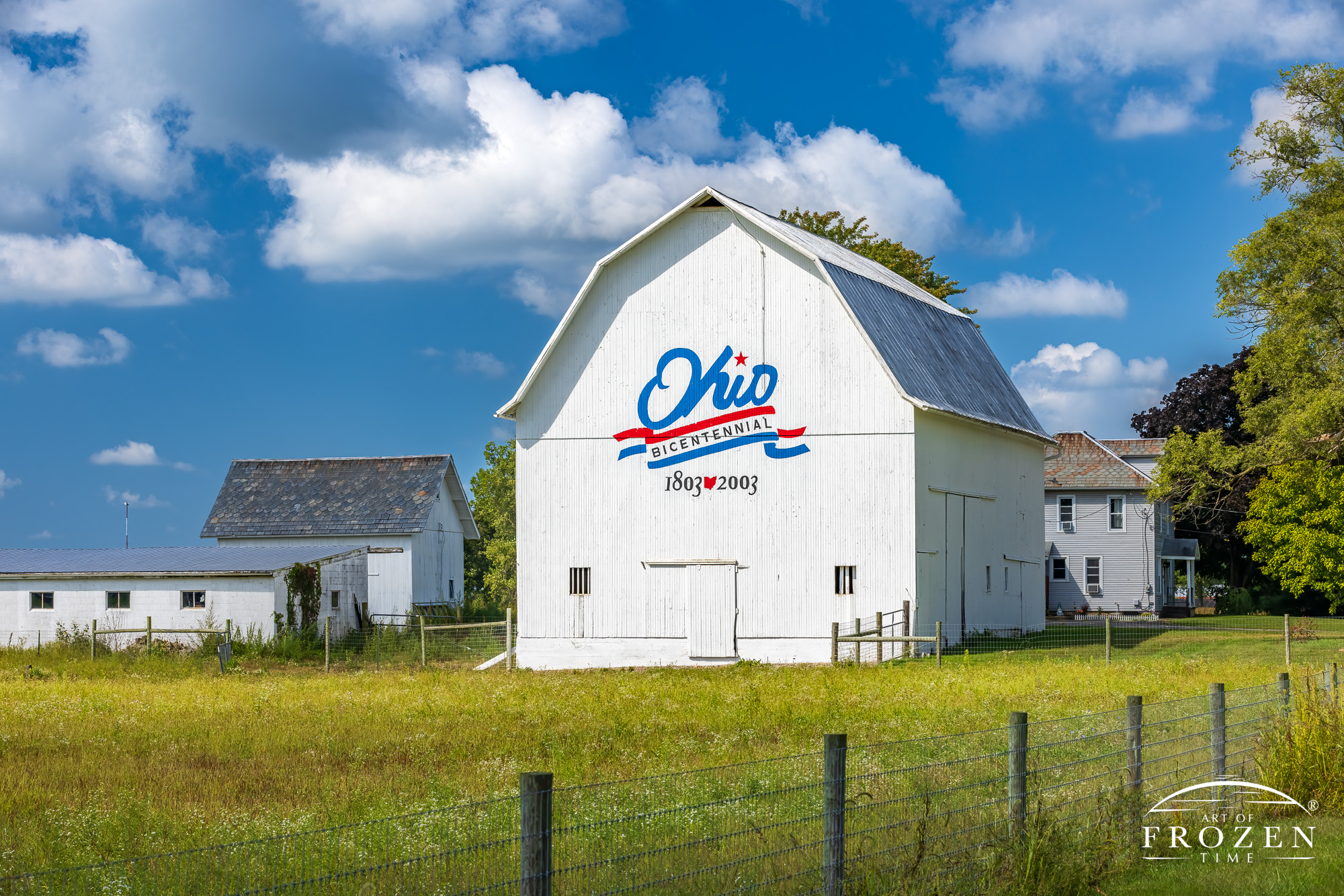 An Ohio art print featuring the nostalgic Marion County Ohio Bicentennial Barn. A bright and well maintained barn featuring the Ohio Bicentennial Logo where the sun shines on the barn art as puffy clouds float through the Ohio Sky