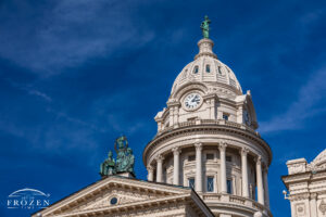 Angled view of the Miami County Courthouse facade in Troy, Ohio, showing Lady Justice, lower bronze figures, sandstone detail, and blue sky.