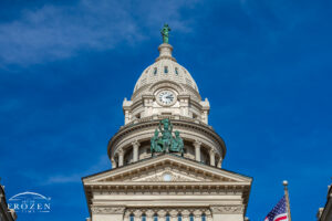 Upper facade of the Miami County Courthouse in Troy, Ohio, showing sandstone architectural detail, Lady Justice, and lower allegorical sculptures against a blue sky.
