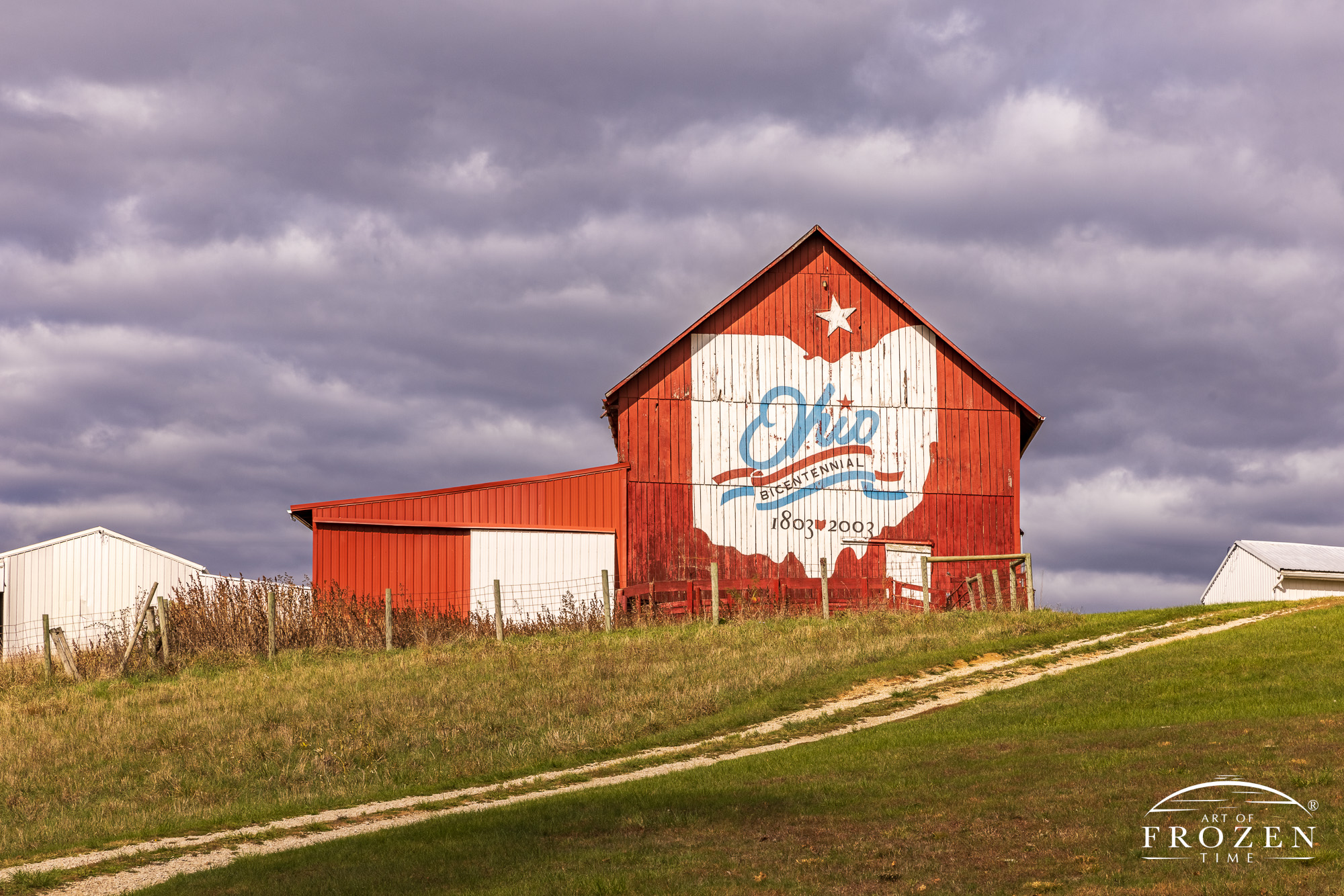 The Morgan County Bicentennial Barn perched on a hillside with its unpaved driveway climbing to the top. The barn and patriotic logo basks in sunlight that broke through the moody clouds.