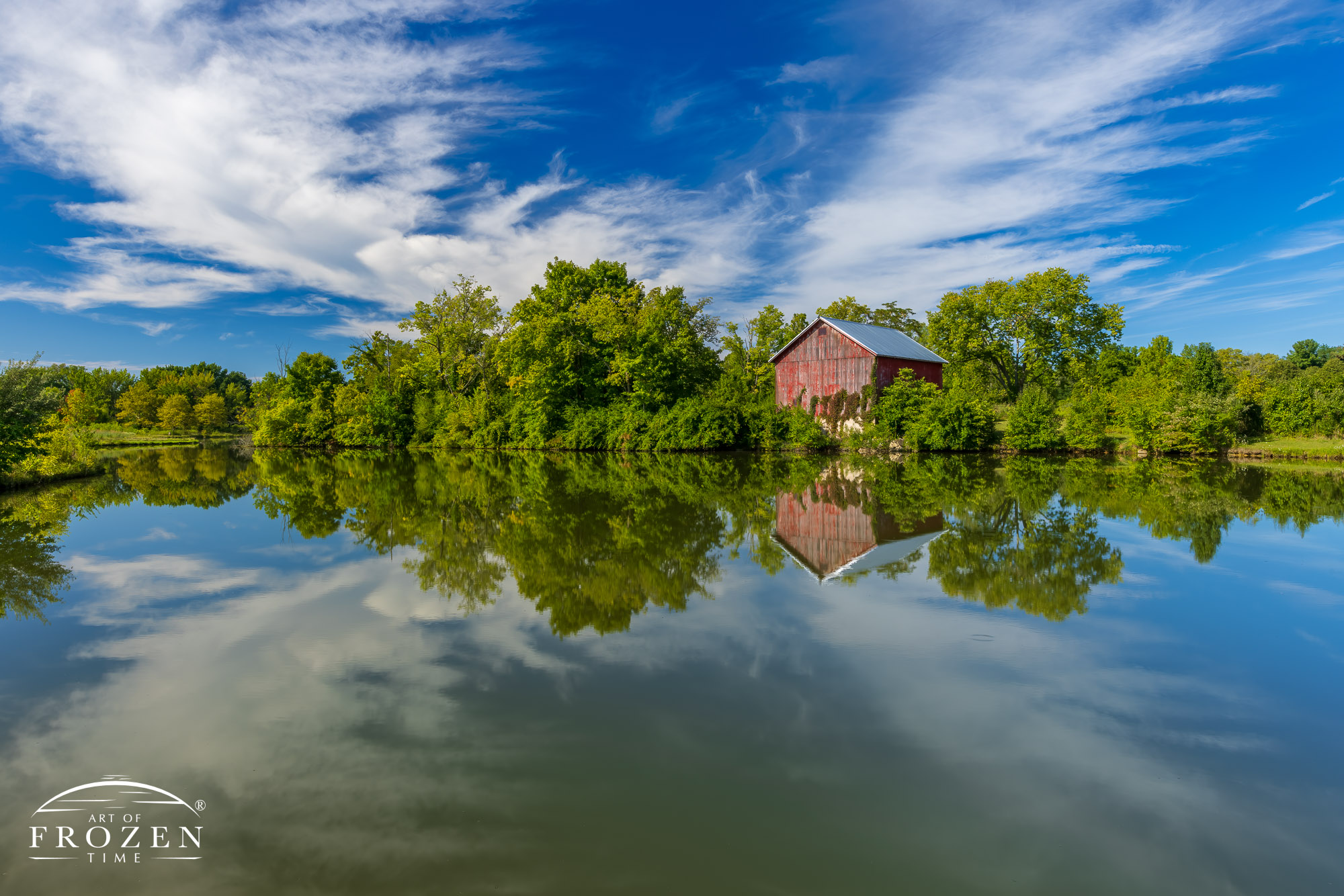 Fine art photography print of an old Ohio barn on a summer morning where the adjacent lake reflects the nostalgic structure as well as the blue sky above.