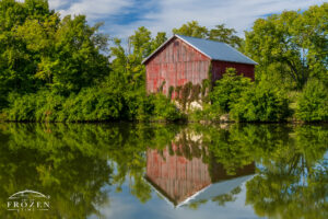 Telephoto view of an old red barn reflected in the calm lake at Elk Creek MetroPark Meadow Ridge near Middletown, Ohio, with surrounding trees and still water.