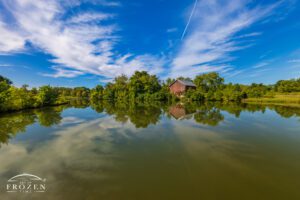 Ultra-wide view of an old red barn reflected in the lake at Elk Creek MetroPark Meadow Ridge near Middletown, Ohio, with blue sky, cirrus clouds, and summer trees.