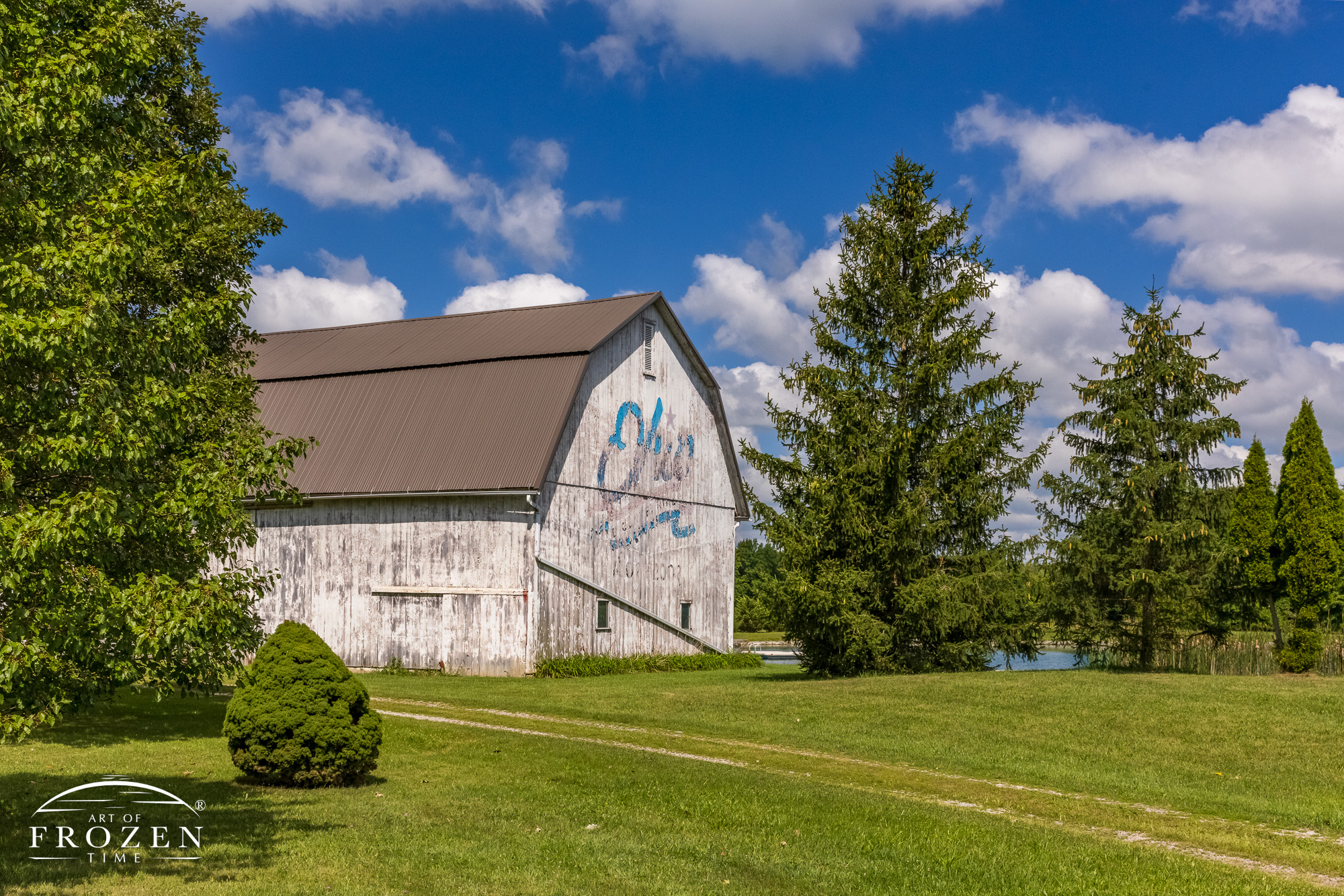 A worn white barn with a single lane drive connecting the barn to the road. The Ohio Bicentennial Logo is readable as cumulus clouds float by in the blue sky.