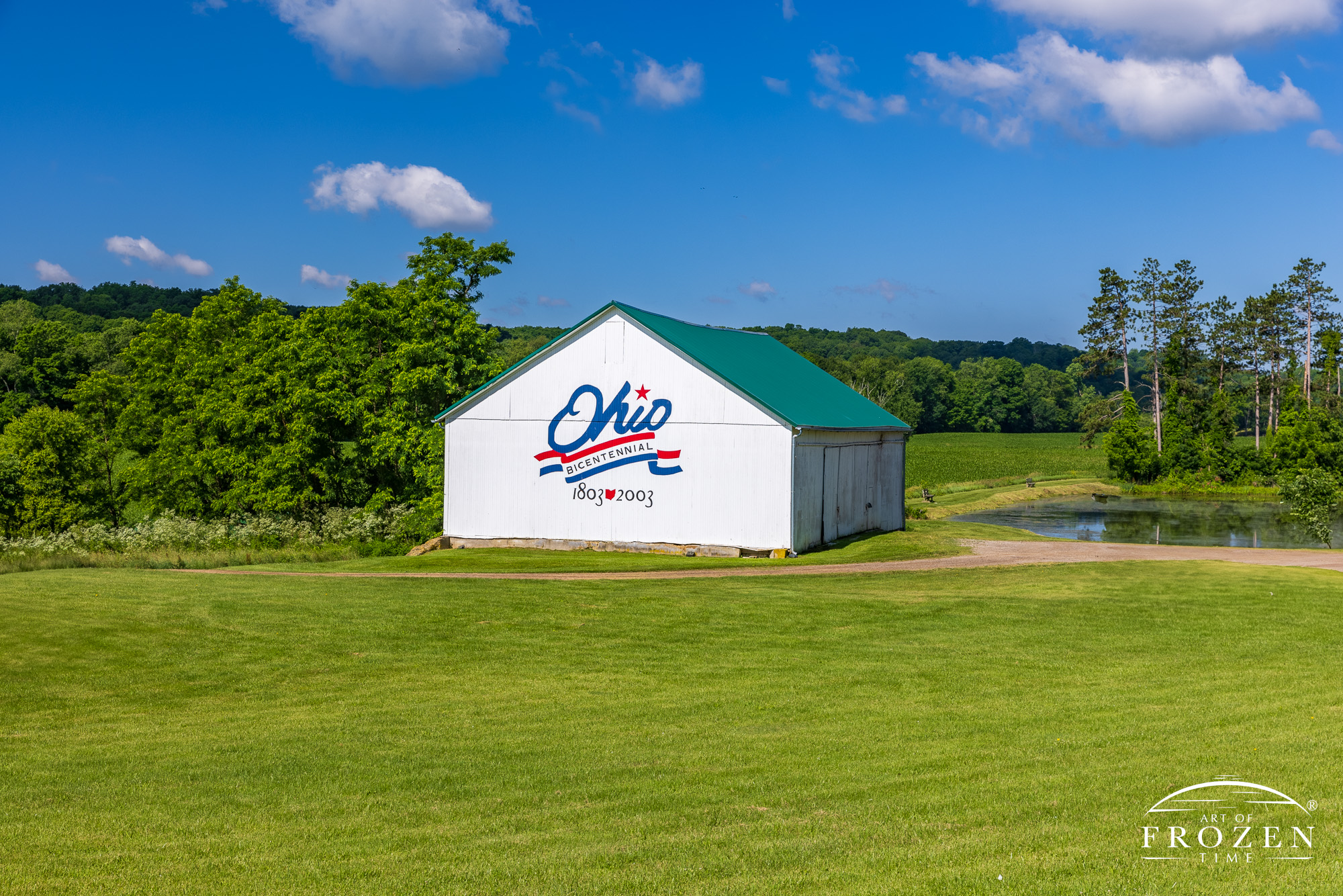 A bright white barn on a pretty June day sitting in a lush valley under blue skies. The barn is painted with the red, white, and blue Ohio Bicentennial Logo