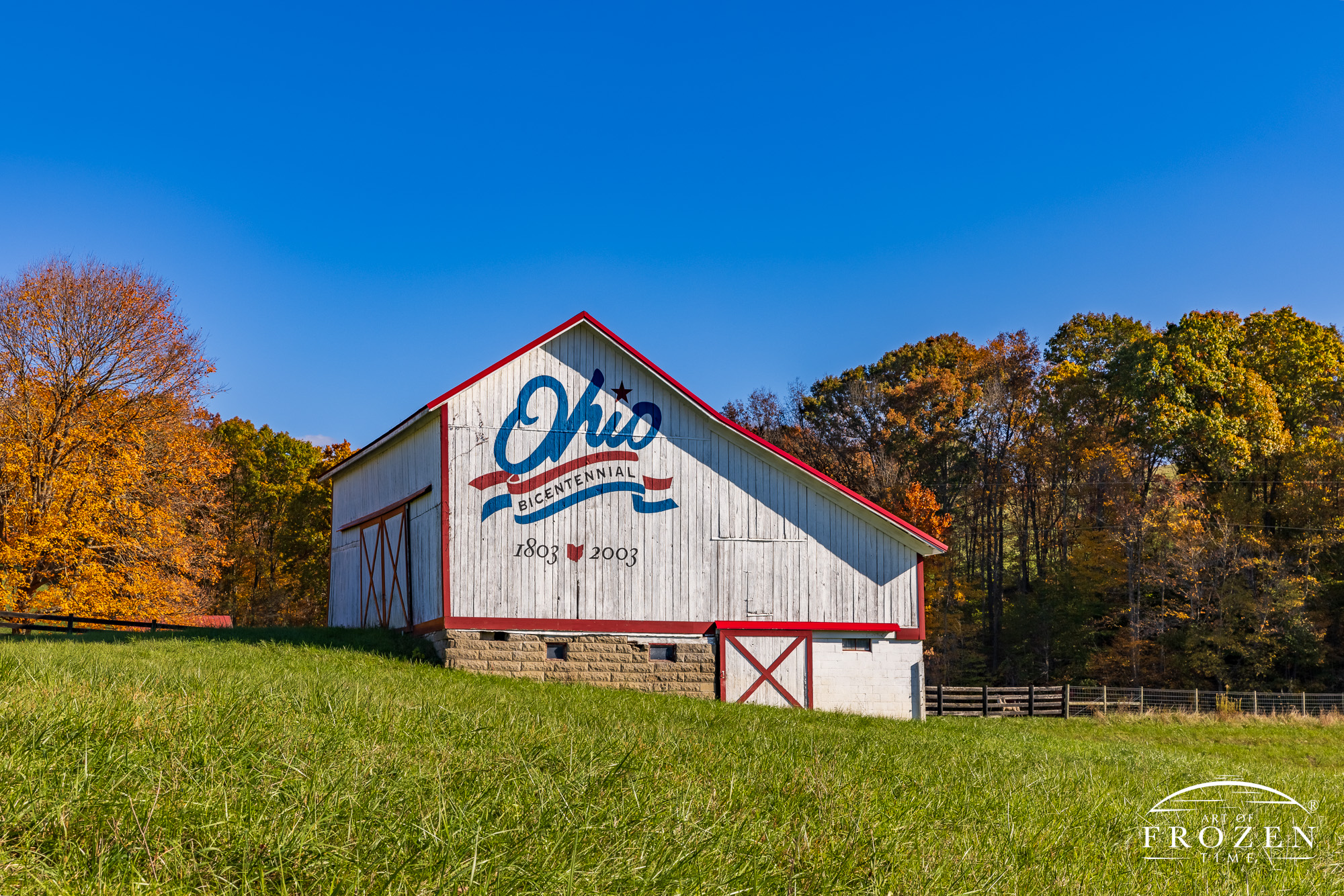 A classic Ohio Bicentennial barn on a bright autumn evening where the white barn contrasts with the red, white and blue logo nestled in an autumn colored hillside.